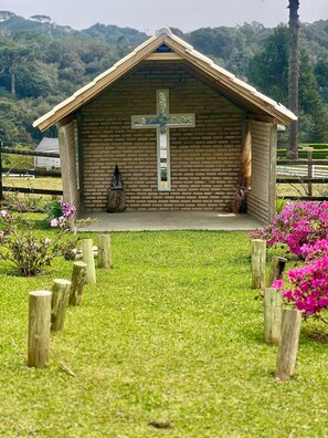 A small brick chapel with a cross, surrounded by a grassy area and wooden posts. - O Chale Encantado (Rancho Queimado)