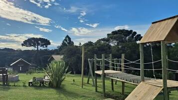 A playground with wooden structures and rope bridges, surrounded by grass and trees.