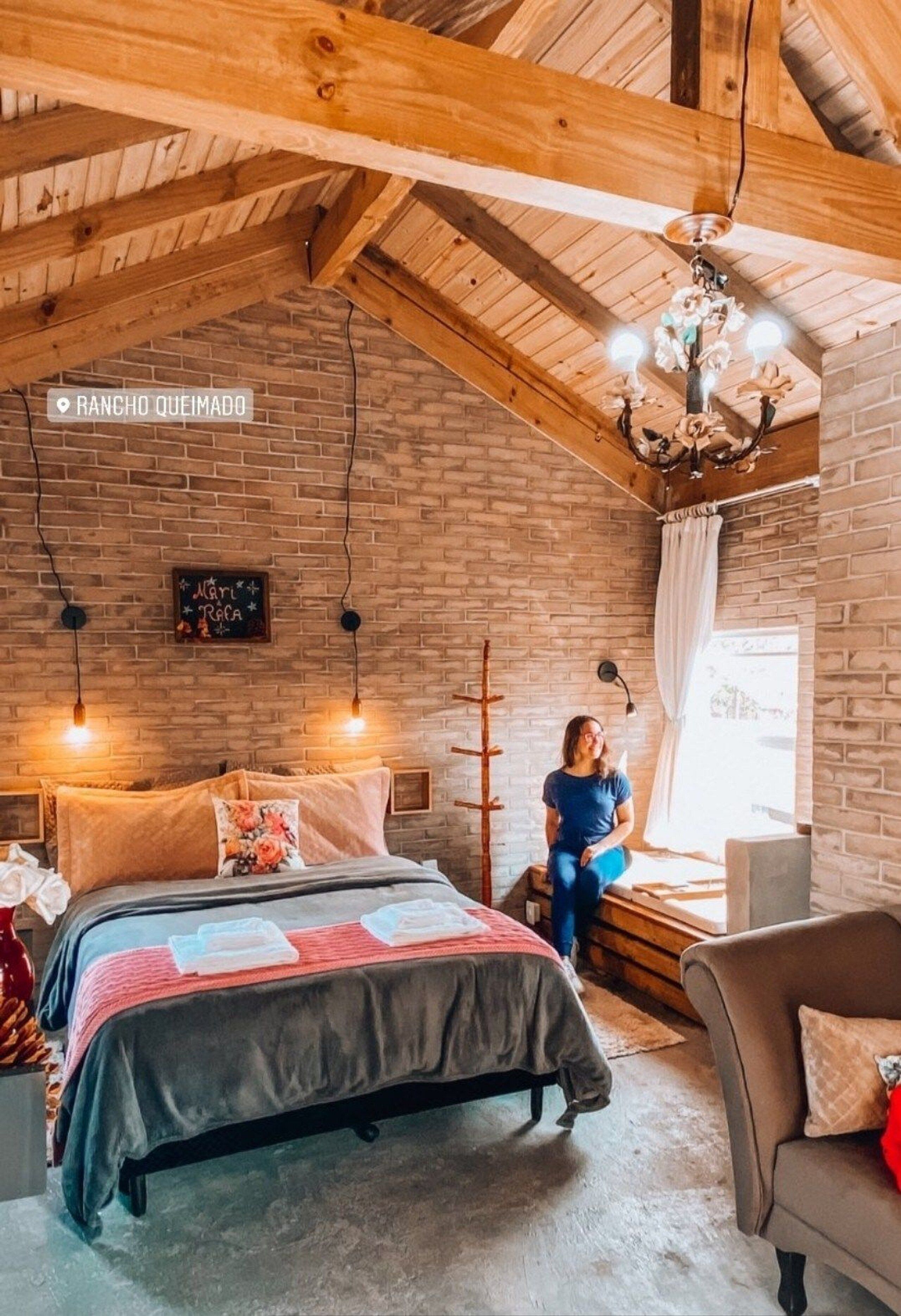 A rustic room with exposed wooden beams, a brick wall, a bed with a floral pillow, a bench, and a chandelier.