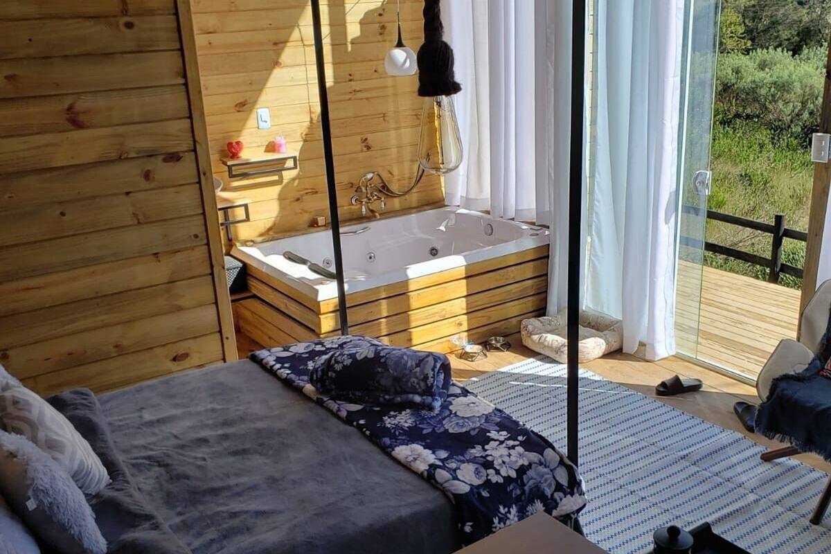 A cozy cabin room with a wooden interior, a bed with a floral duvet, a small wooden table, and a glass-enclosed balcony with a view of greenery.