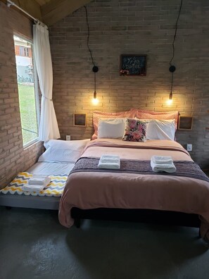 A bedroom with a bed, a small sofa, a window with curtains, and a brick wall. - O Chale Encantado (Rancho Queimado)