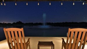 Une terrasse en bois avec deux chaises et une petite table, donnant sur une étendue d’eau agrémentée d’une fontaine et surplombant un ciel nocturne.