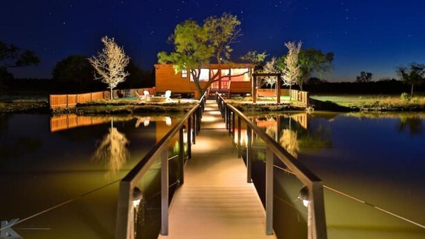 Un pont en bois mène à un bâtiment moderne doté d’une façade en verre, entouré d’arbres et bordé par une étendue d’eau paisible.