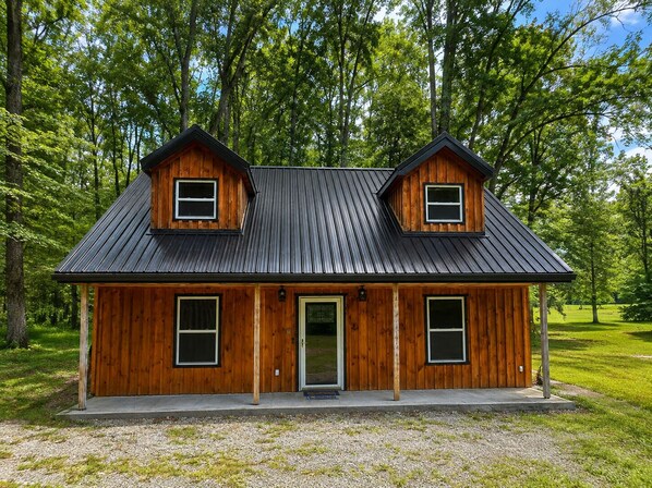 Una cabaña de madera con techo de metal, dos ventanas en la fachada y una puerta.