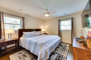 Ein ordentlich eingerichtetes Schlafzimmer mit Bett, Nachttischen, Schreibtisch, Deckenventilator und großen Fenstern mit Vorhängen.