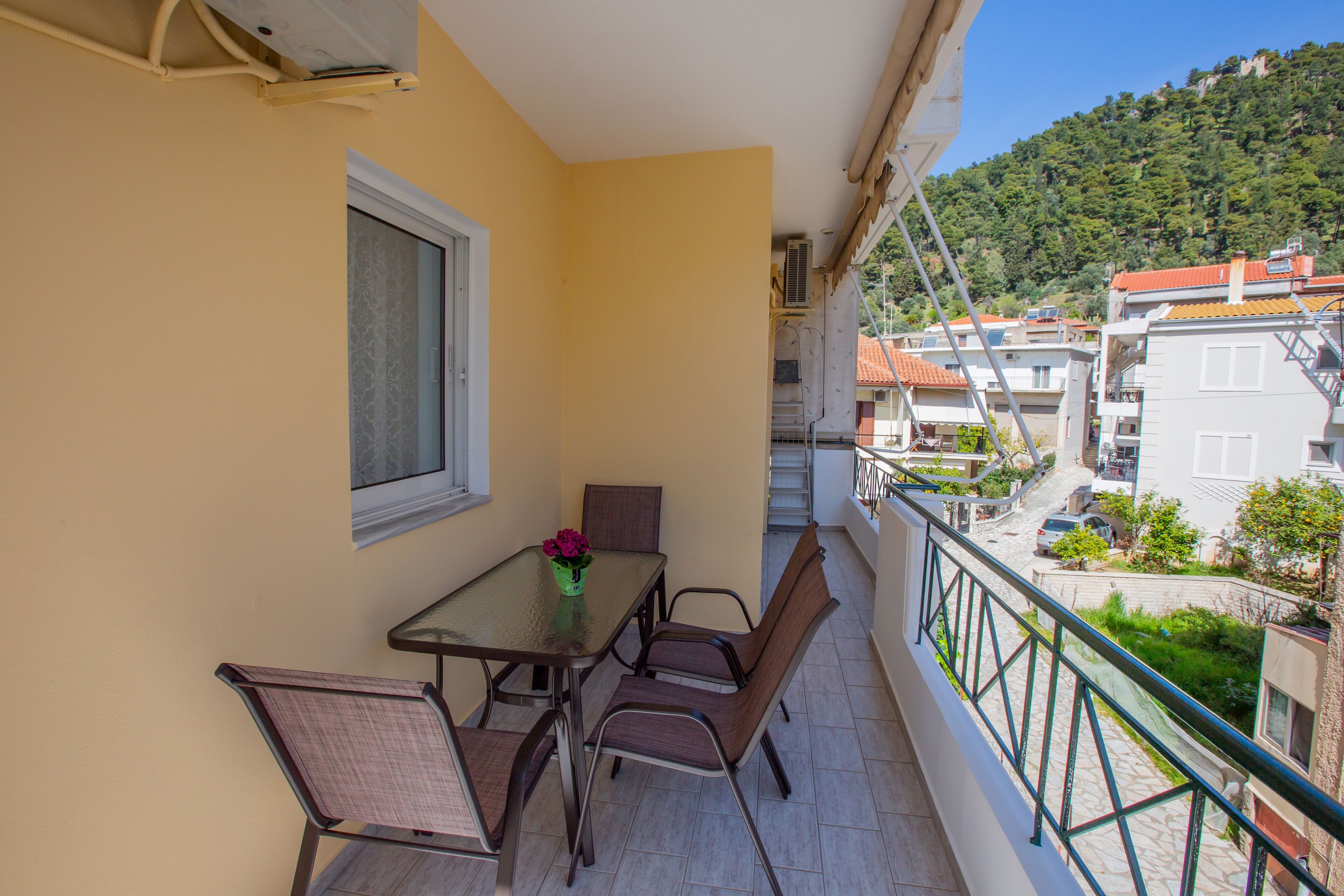 A balcony with a table and chairs, a window, and a view of buildings and a hill.