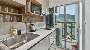 A kitchen with a stainless steel sink, a microwave, and a kettle. There is a balcony with a view of a hilly landscape. - Dcondo Kathu - Top Floor 1 Bdr Condo with shared Pool (Kathu)