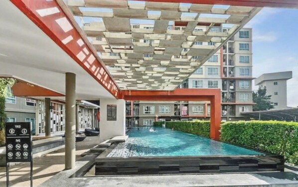 Una piscina con una toldería roja y blanca, rodeada de vegetación y contigua a un edificio de varios pisos.