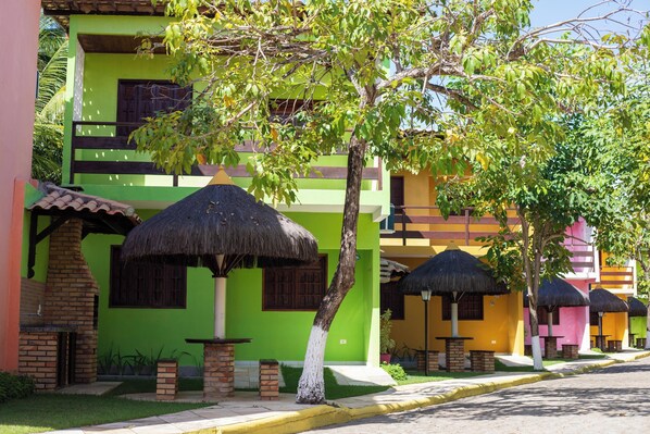 A row of colorful buildings with thatched-roof structures, a paved street, and a clear sky. - PRIVÊ RESIDENCIAL MARAGOGI (Maragogi)