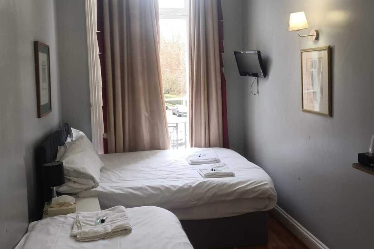 A hotel room with two beds, a window with curtains, a wall-mounted TV, and a small table with a lamp.