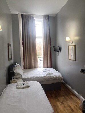 A hotel room with two beds, a window with curtains, a wall-mounted TV, and a small table with a lamp. - Coronation Hotel (London)