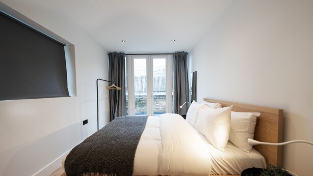 A modern bedroom with a bed, a bedside table with a plant, a hanging light, and a view of the city through large windows.