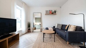 A modern living room with a flat-screen TV on a wooden stand, a dark sofa with multiple cushions, a small round coffee table, and a floor lamp.