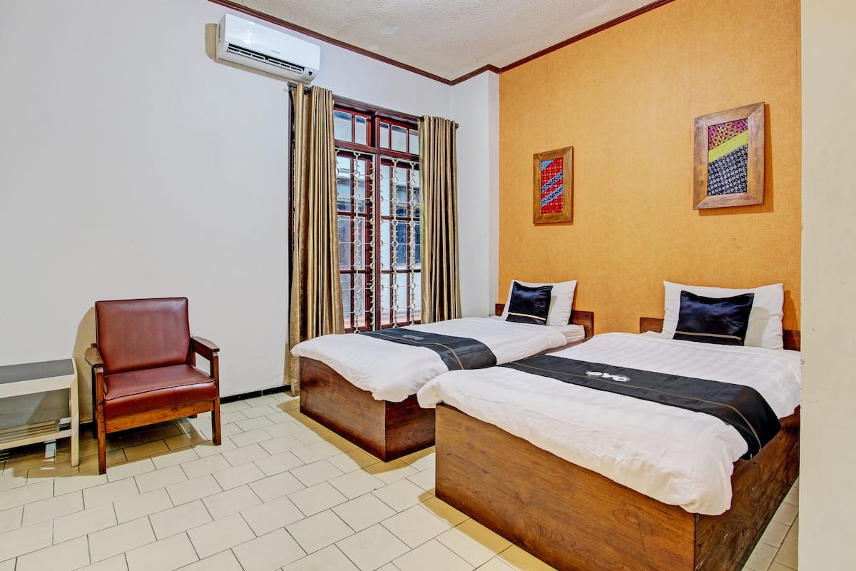 A hotel room with two wooden beds, a leather chair, a window with curtains, and two framed artworks on the wall.