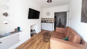 A modern living room with a flat-screen TV, a sofa, a coffee table, and a kitchen area in the background. - Beautiful 7 bedroom Victorian home in Lancashire (Morecambe)