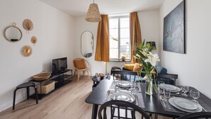 Ein moderner Essbereich mit einem schwarzen Tisch, gedeckt für vier Personen, einem Fernseher auf einem schwarzen Lowboard und einem großen Fenster mit Vorhängen.