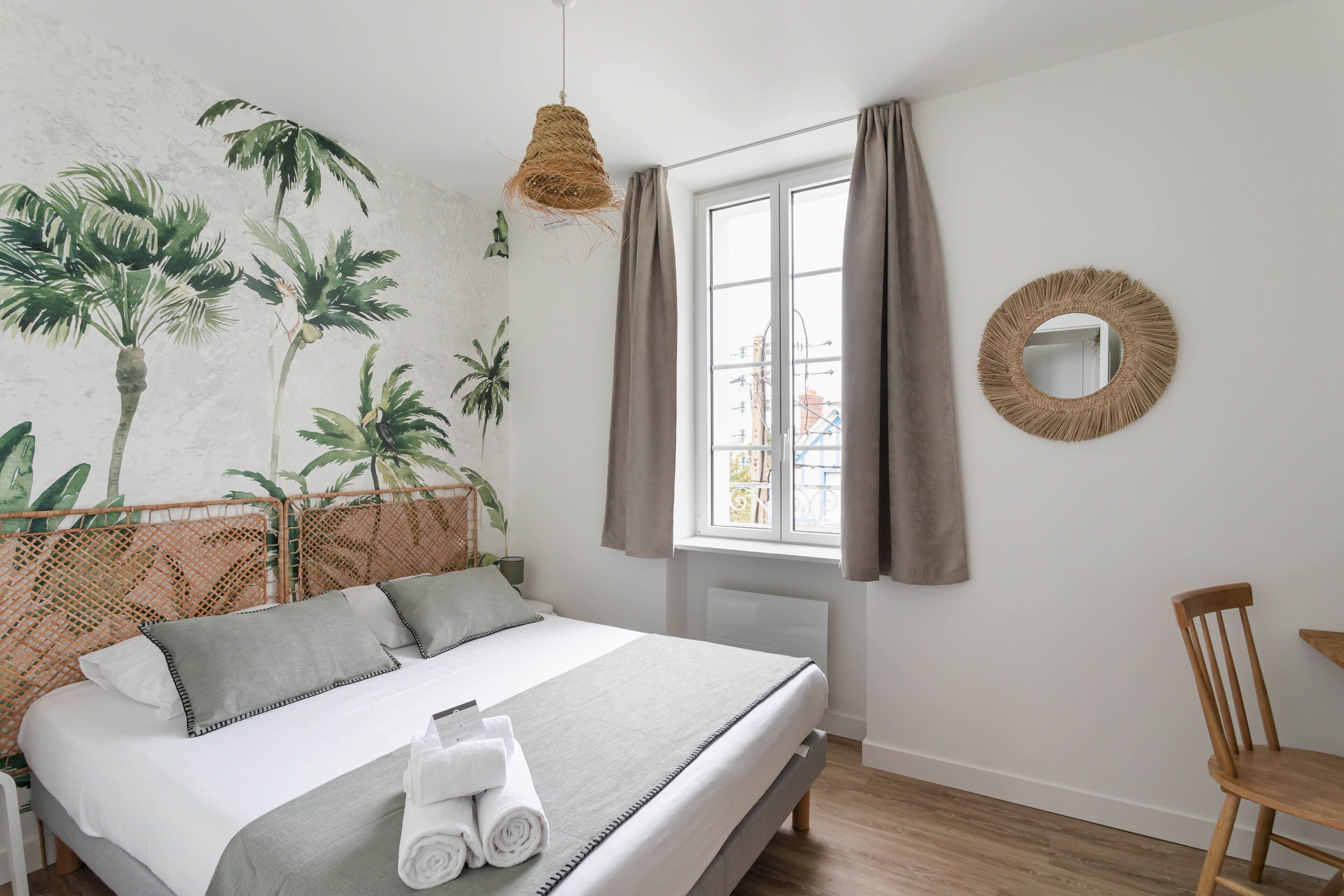 A bedroom with a bed, a chair, a window with curtains, a wall-mounted mirror, and a wall mural of palm trees.