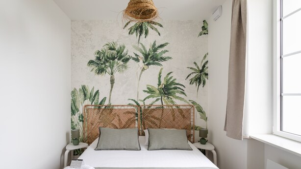 Ein Schlafzimmer mit einem Bett, einem gewebten Kopfteil, einem Fenster mit Vorhängen und einer Wand mit einem tropischen Pflanzenwandbild.