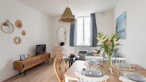 Ein moderner Essbereich mit einem Holztisch, gedeckt für vier Personen, einem Fernseher auf einer Holzkonsole und einem großen Fenster mit Vorhängen.