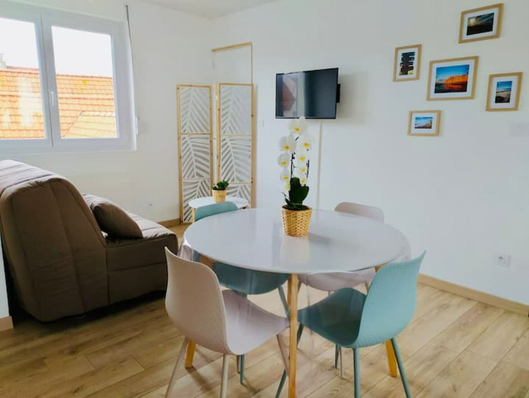 Un salon moderne avec une table ronde, quatre chaises, un canap é, un téléviseur fixé au mur et une fenêtre donnant sur un immeuble.
