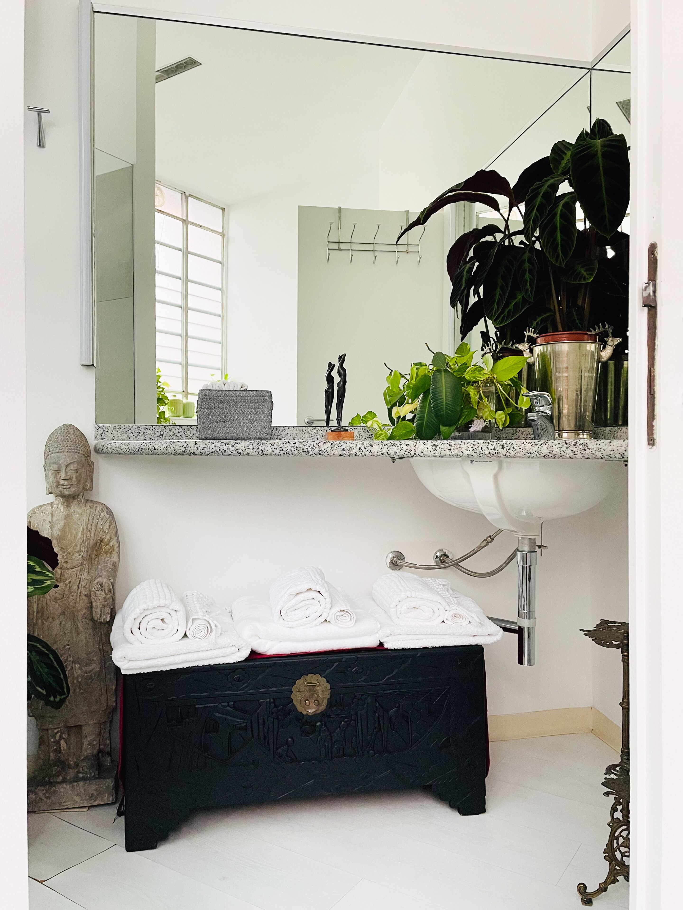 Un bagno con uno specchio grande, un lavandino, una statua, un porta asciugamani e una pianta.