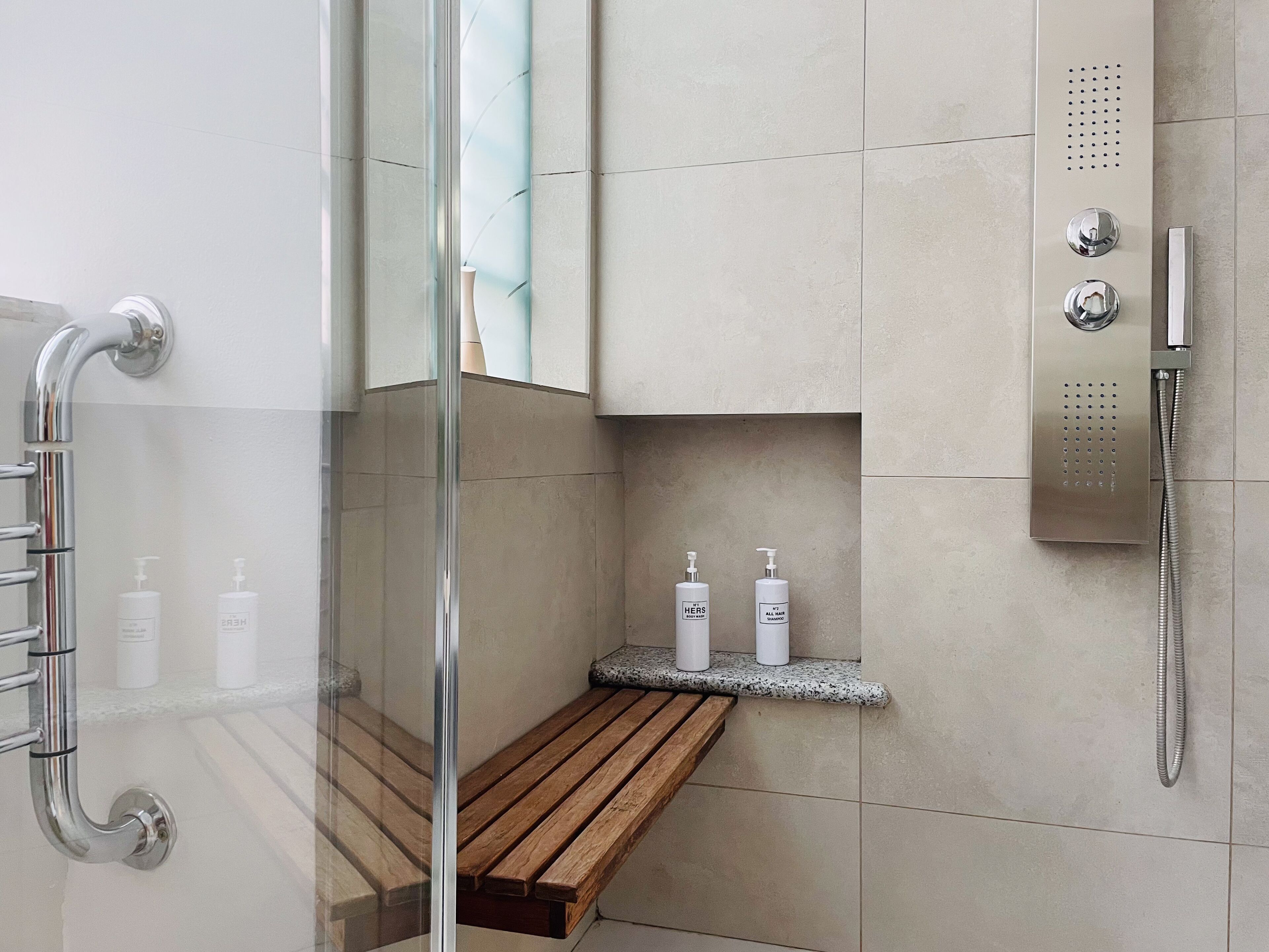 A modern bathroom with a glass shower enclosure, a wooden bench, and a shelf with toiletries.