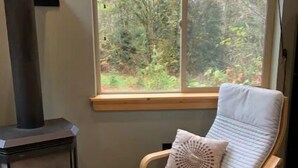 A room with a wood-burning stove, a window with a view of trees, and a wooden chair with a cushion. - The Glacier Nook (Deming)