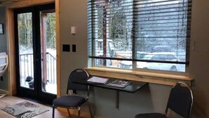 A room with a wooden door, a window with blinds, a small table with a chair, and a rug on the floor. - The Glacier Nook (Deming)