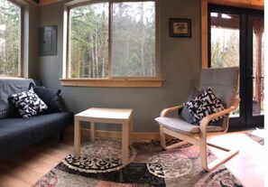 A cozy living room with a dark blue sofa, a wooden coffee table, and a grey armchair with patterned cushions. Large windows offer a view of a forested area. - The Glacier Nook (Deming)