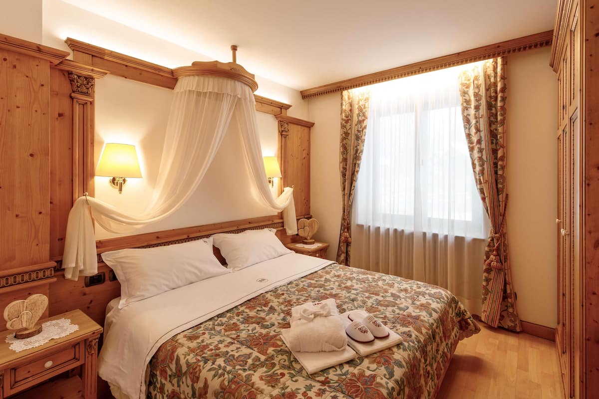 Una habitación de hotel con cama con dosel, cabecero de madera y dosel. Hay dos mesitas de noche con lámparas y una mesita pequeña con un teléfono. La habitación tiene ropa de cama con estampado floral y una ventana con cortinas.