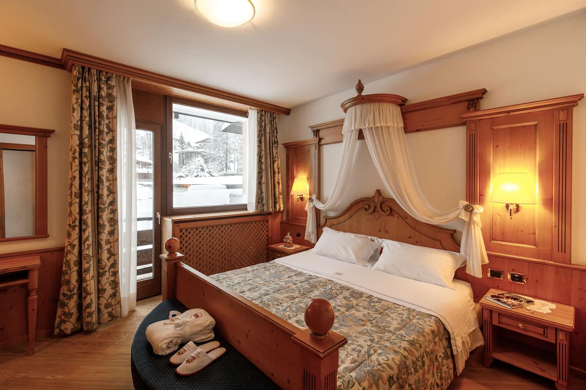 Habitación de hotel con una cama de dos columnas de madera, una ventana con cortinas, una mesita de noche con una lámpara y una pequeña mesa con un libro y un objeto decorativo.