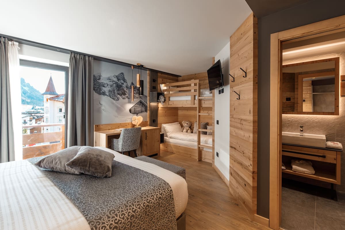 Una habitación de hotel moderna con una cama grande, un escritorio, una cama nido y un baño con espejo y lavamanos.