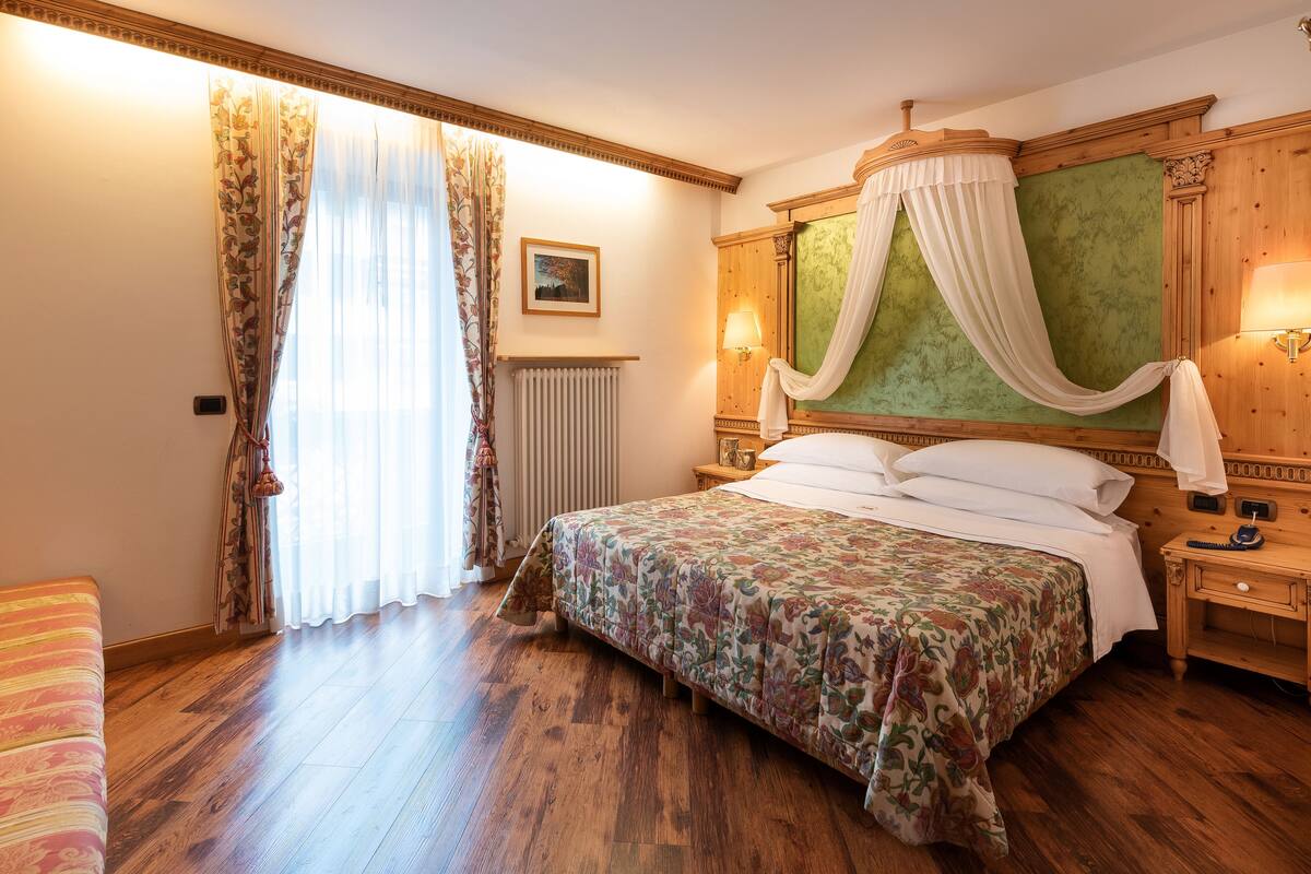 Un dormitorio con una cama grande, suelo de madera, un radiador, una mesita de noche y una ventana con cortinas.
