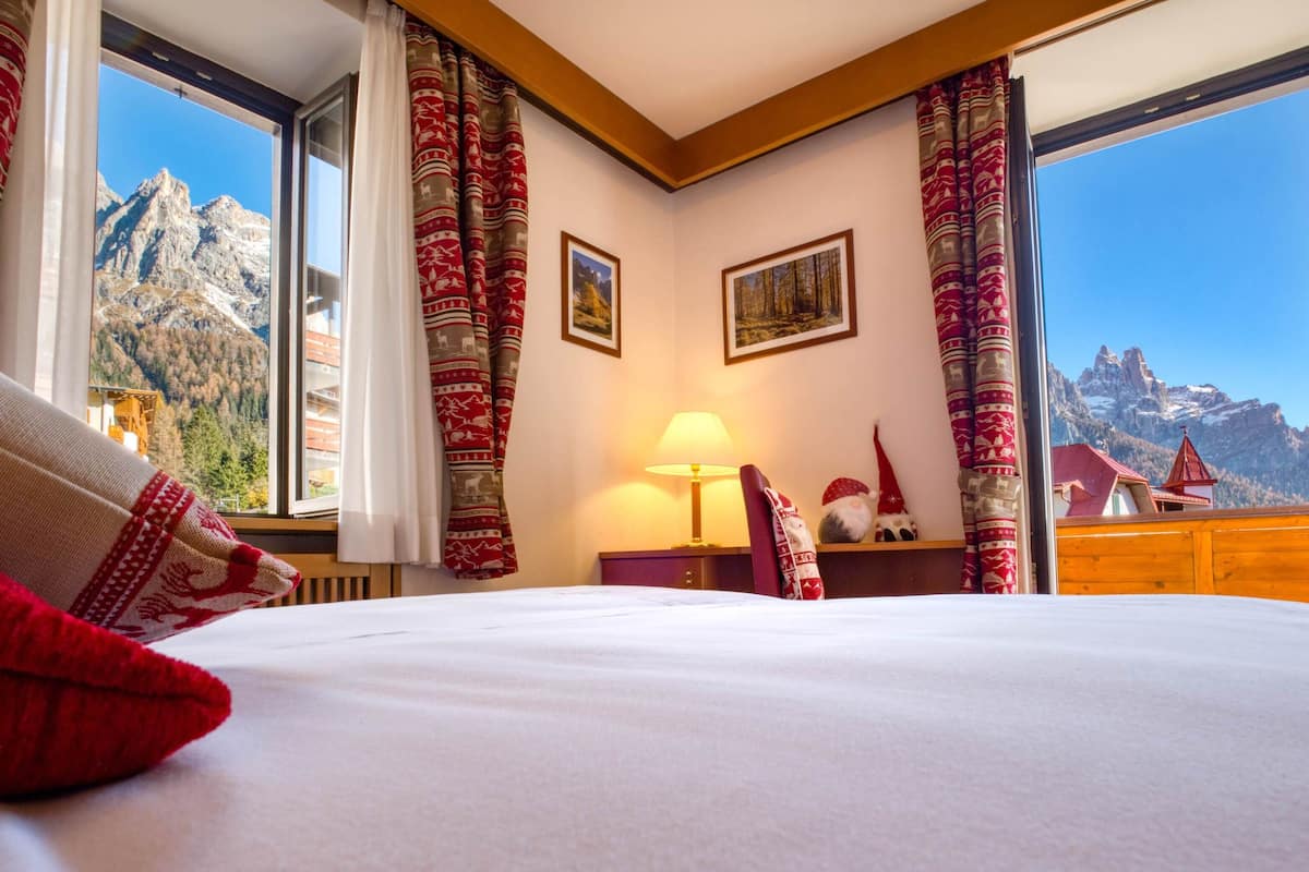 Habitación de hotel con cama, mesita de noche, lámpara y vistas a montañas a través de amplias ventanas.
