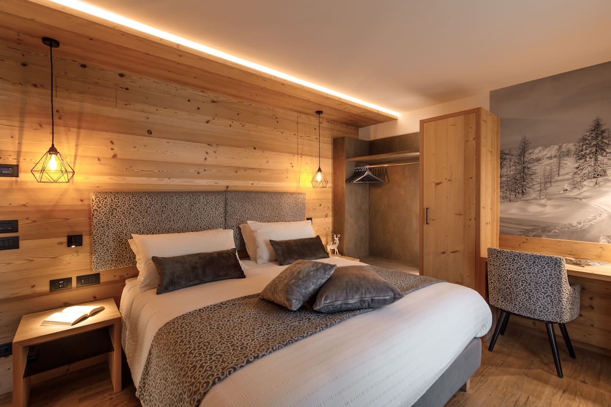 Un dormitorio moderno con techo de madera, una cama con ropa de cama estampada, una mesita de noche, una silla y un cuadro de un paisaje nevado.