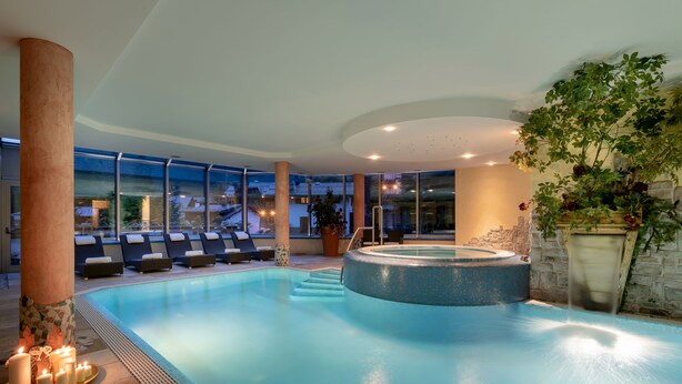 An indoor swimming pool with lounge chairs, a hot tub, and a large window offering a view of the outside.