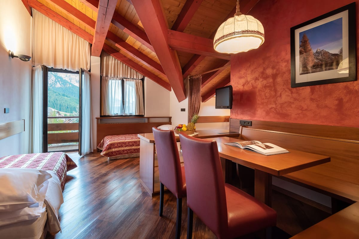 Habitación de hotel con techo de madera, comedor con mesa y sillas, cama con colcha estampada en rojo y blanco, y vista a la montaña a través de la ventana.