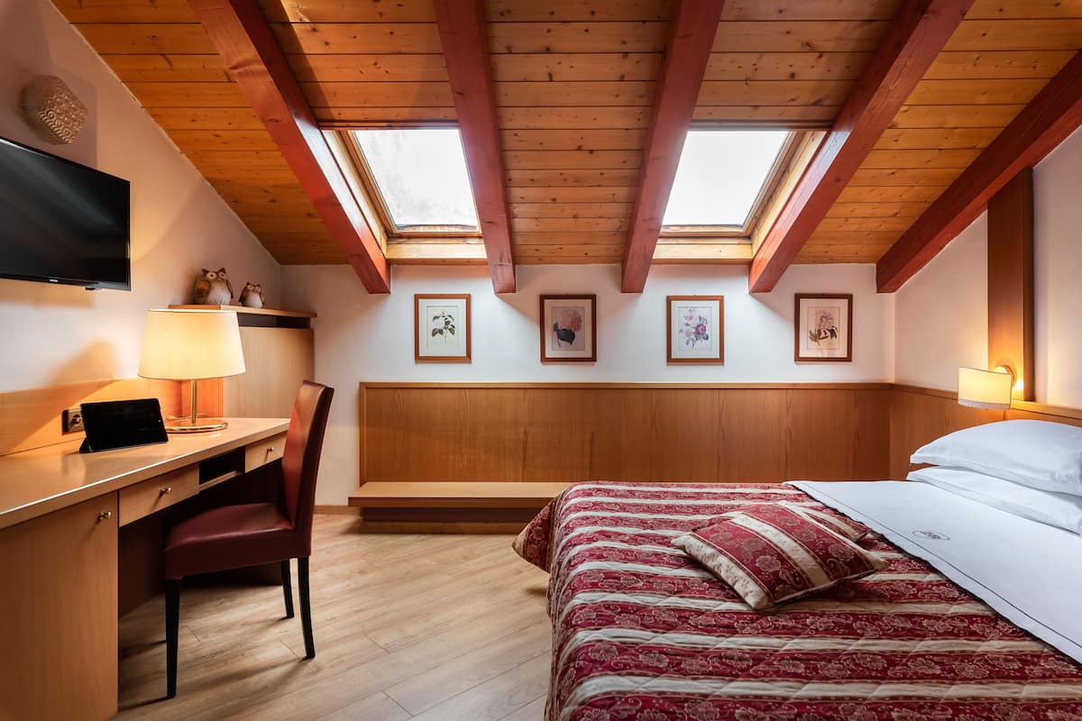 Un dormitorio con escritorio de madera, silla y cama. Hay tragaluces en el techo, un televisor y cuadros en la pared.