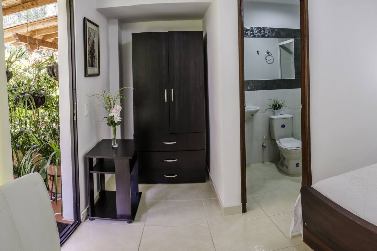 Un dormitorio con cama, mesitas de noche, armario, baño con inodoro y lavabo, y una puerta que da a un balcón.
