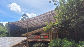A covered outdoor area with a wooden deck and seating, surrounded by lush greenery and a road.