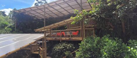 A covered outdoor area with a wooden deck and seating, surrounded by lush greenery and a road.