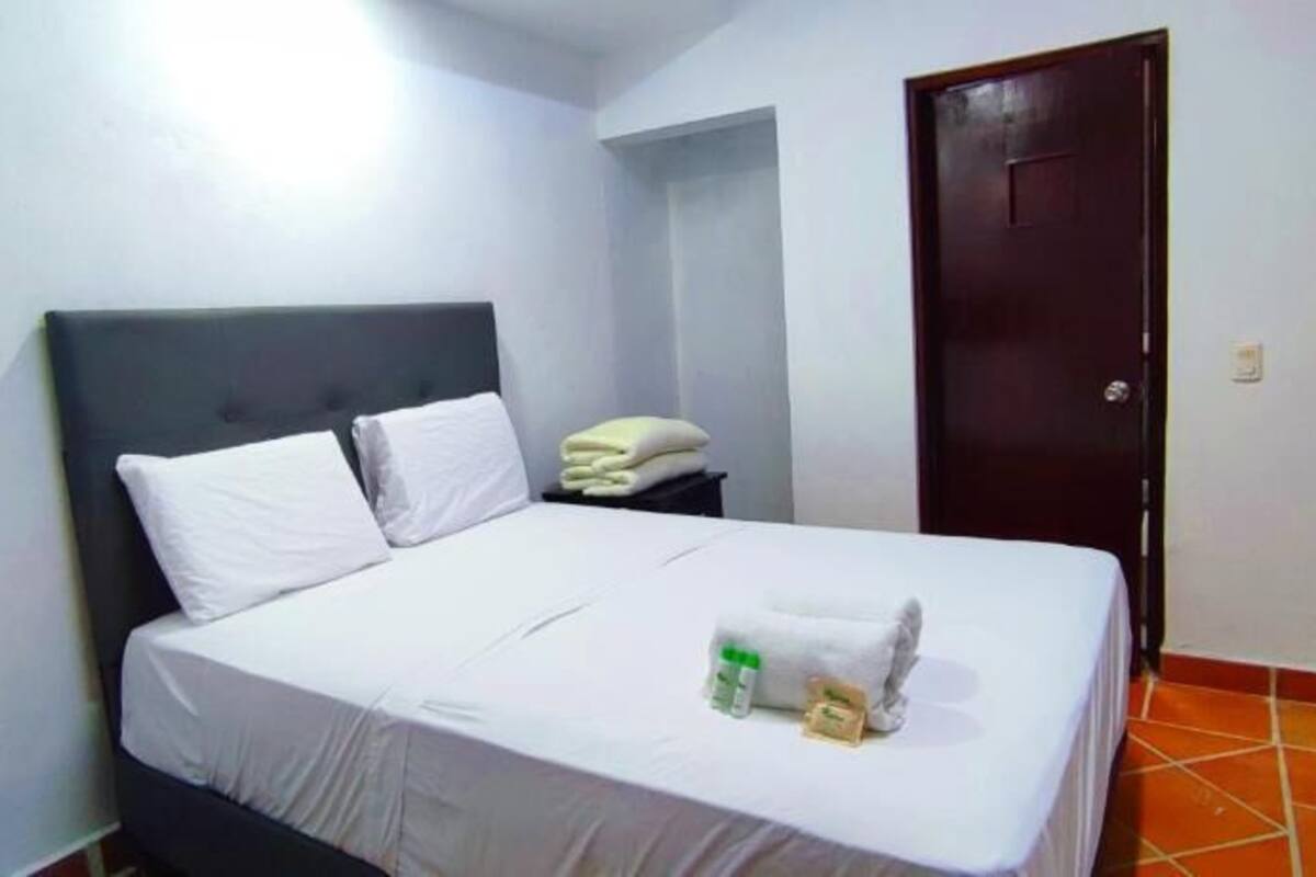 Una cama bien hecha con sábanas blancas, una mesita de noche con una toalla doblada y una puerta a la derecha.