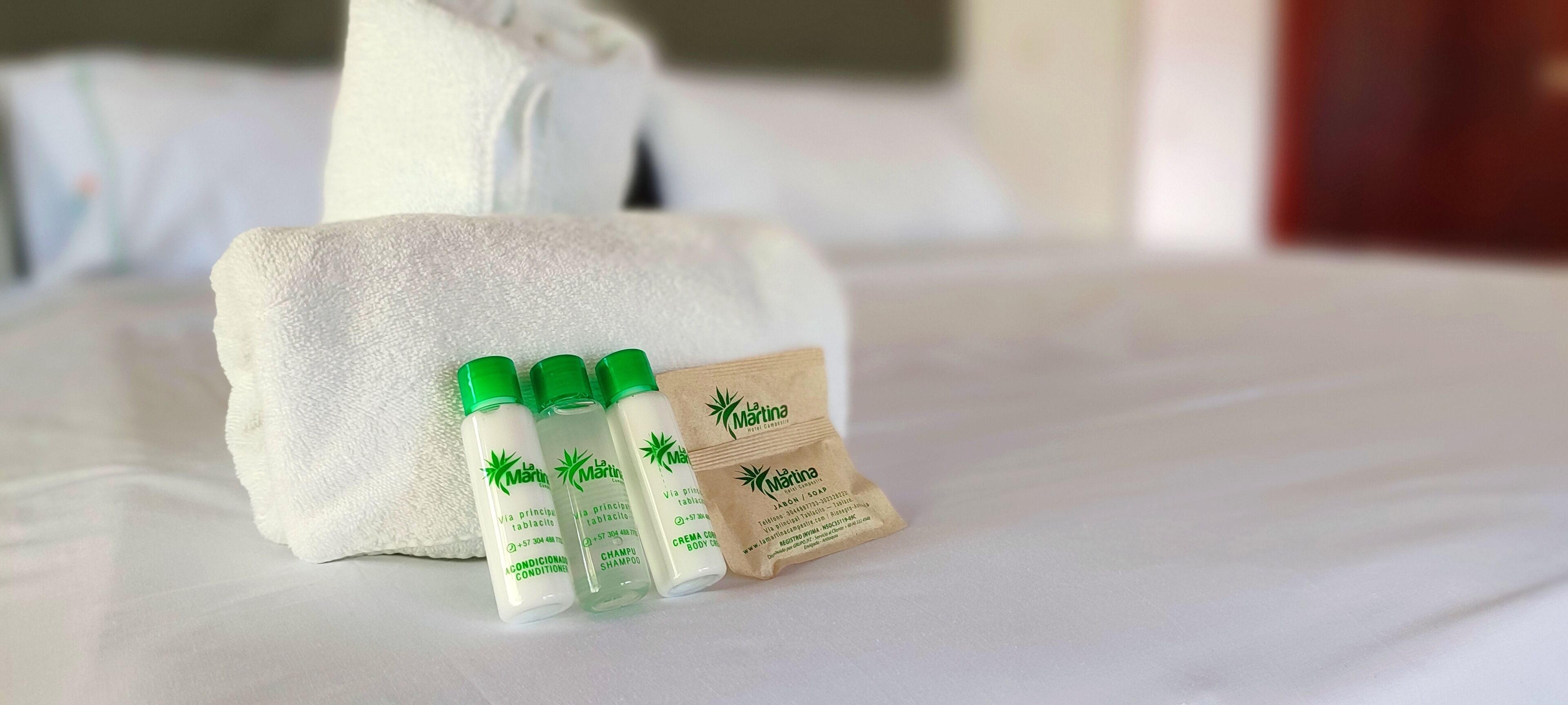 A neatly made bed with a white towel and three bottles of La Martinia shampoo and conditioner, and a small brown packet of body cream.