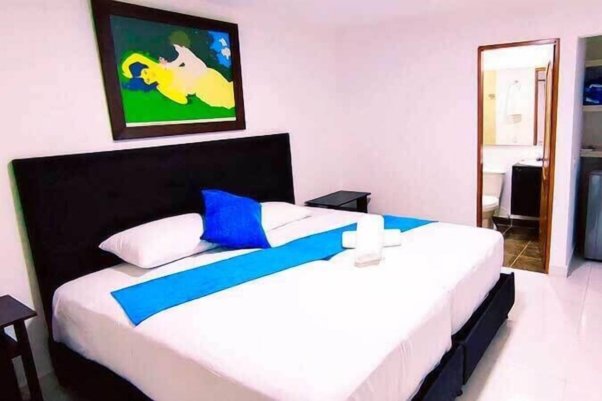 Una habitación de hotel con cama, mesita de noche, un cuadro y baño visible a través de una puerta abierta.