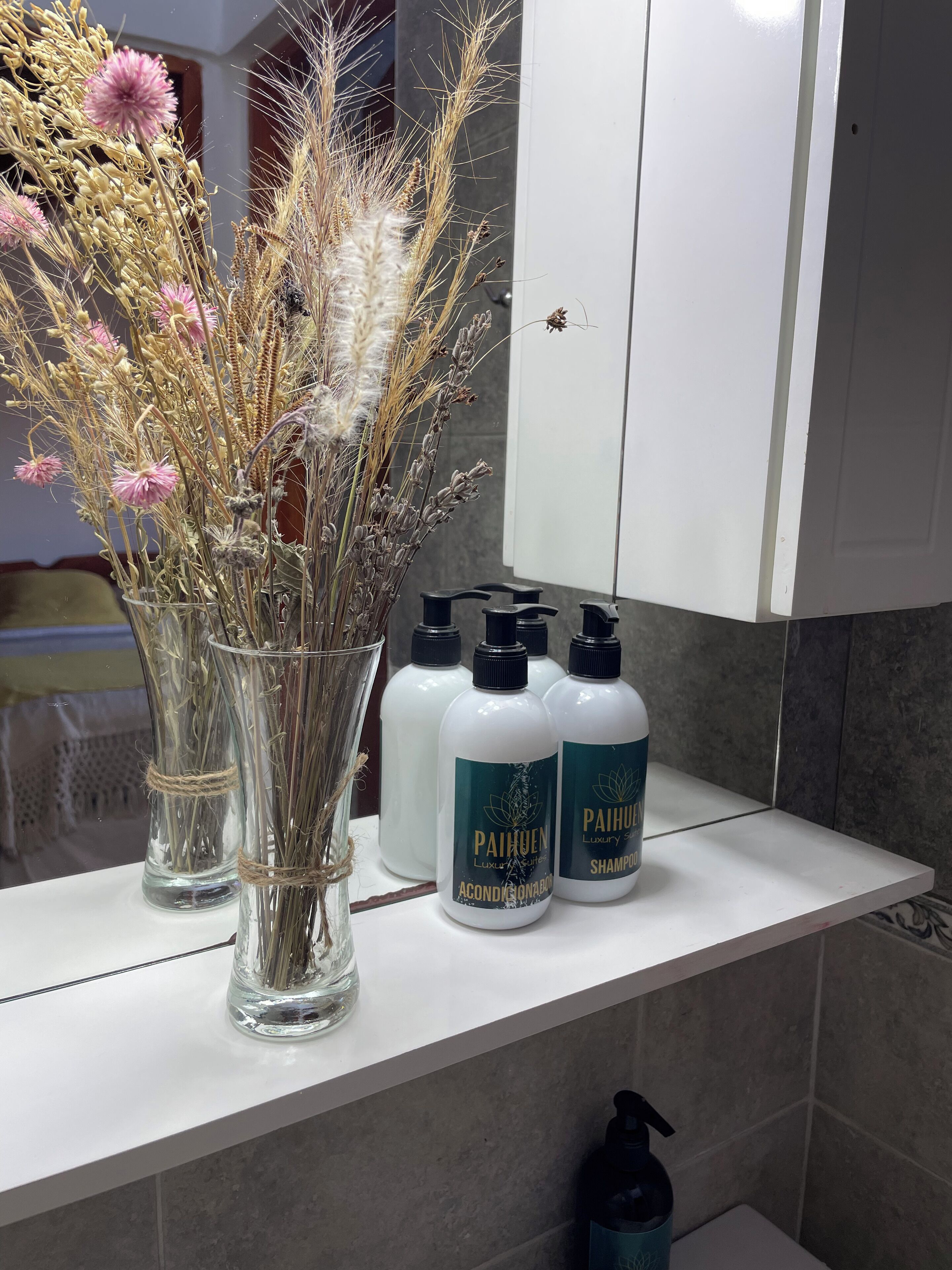 Un meuble-lavabo de salle de bain orné d’un vase de fleurs séchées, de deux bouteilles de shampoing Paihun et d’un flacon d’après-shampoing.