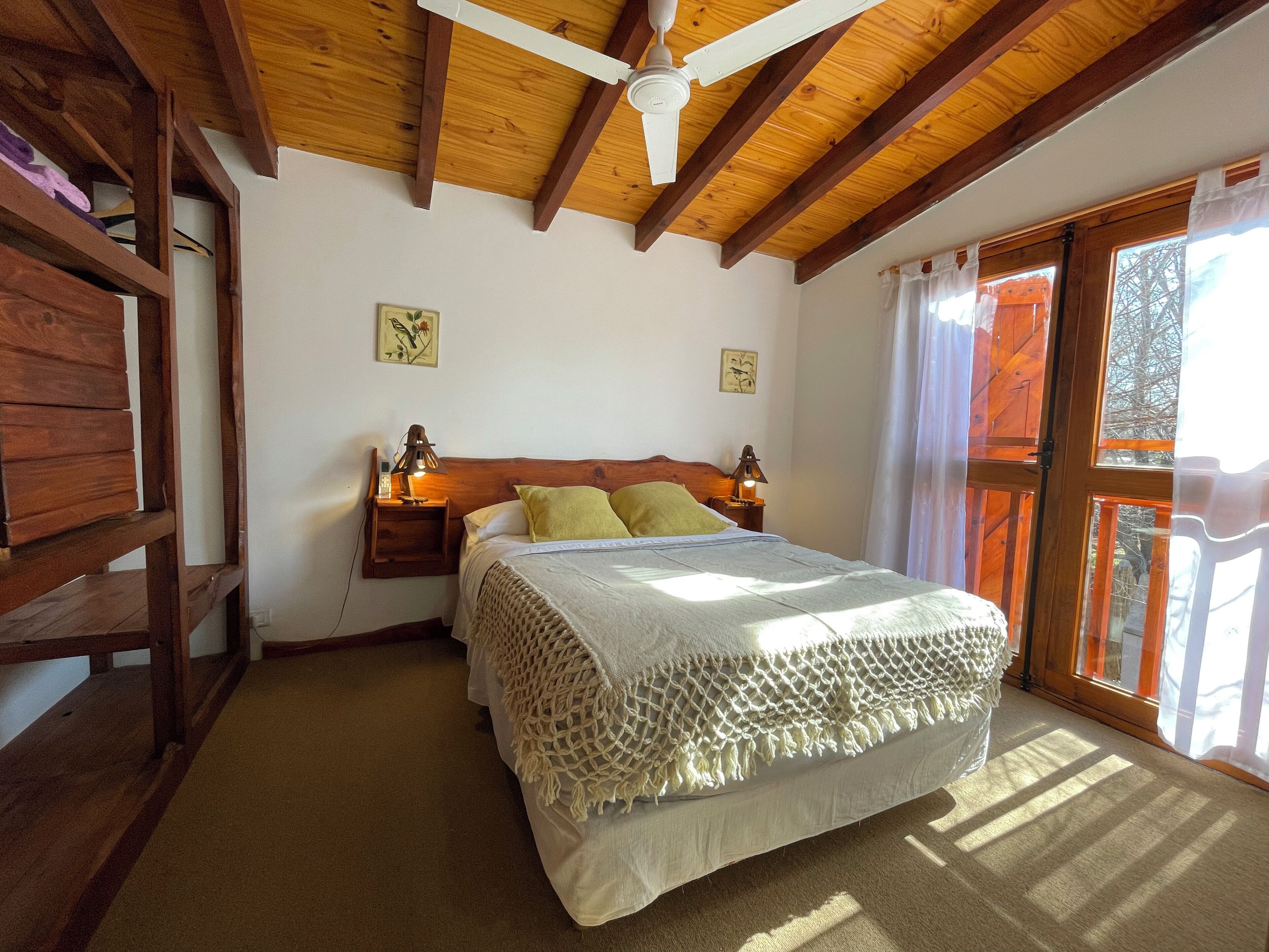 Un dormitorio con techo de madera, cama con colcha blanca y almohadas verdes, dos lámparas de noche y una ventana con cortinas.