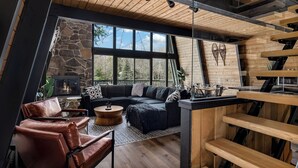 Un interno moderno con un camino in pietra, un divano angolare nero in tessuto, poltrone in pelle e un soffitto in legno.