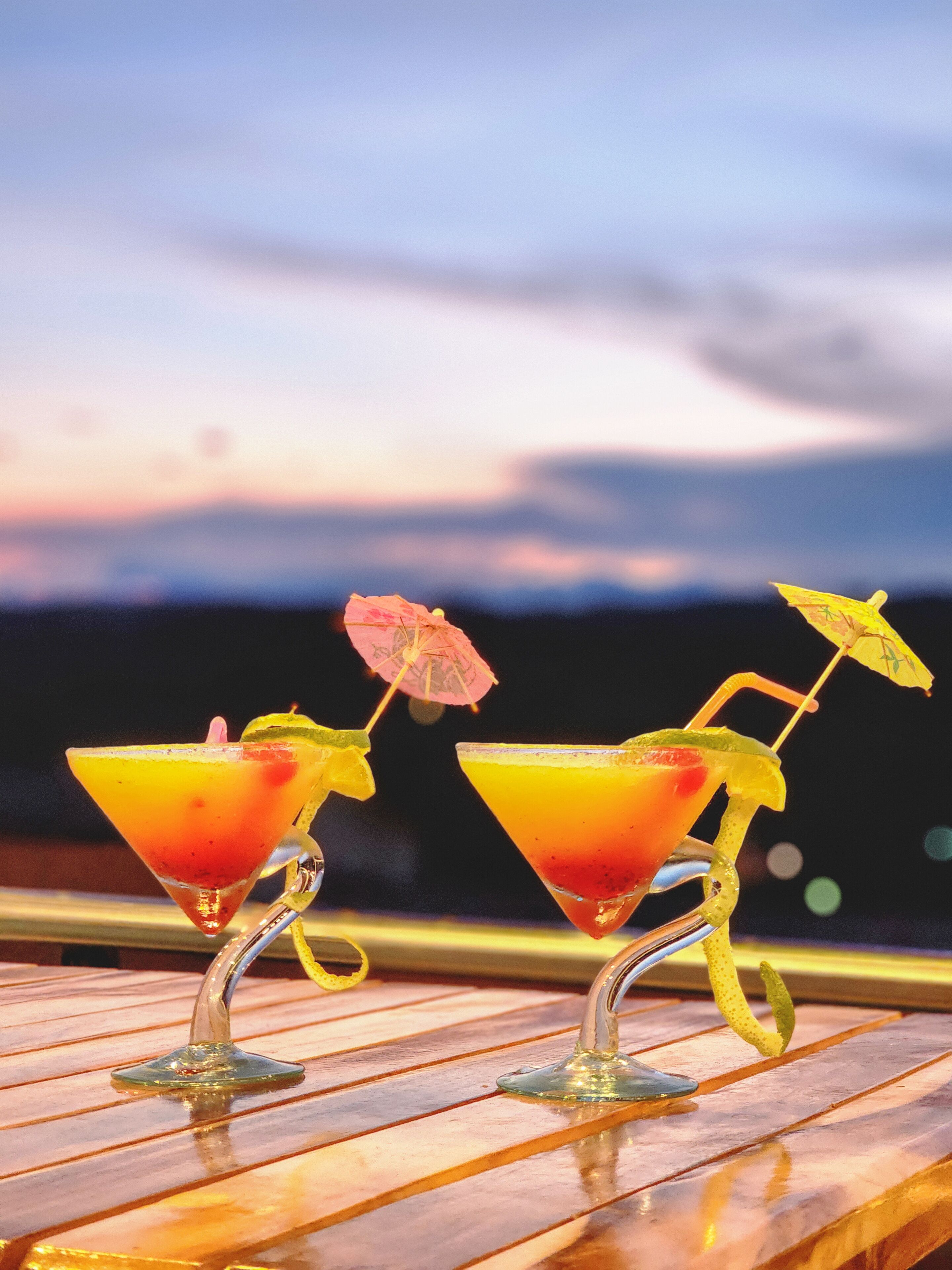 Two martini glasses with umbrellas on a wooden table against a sunset backdrop.