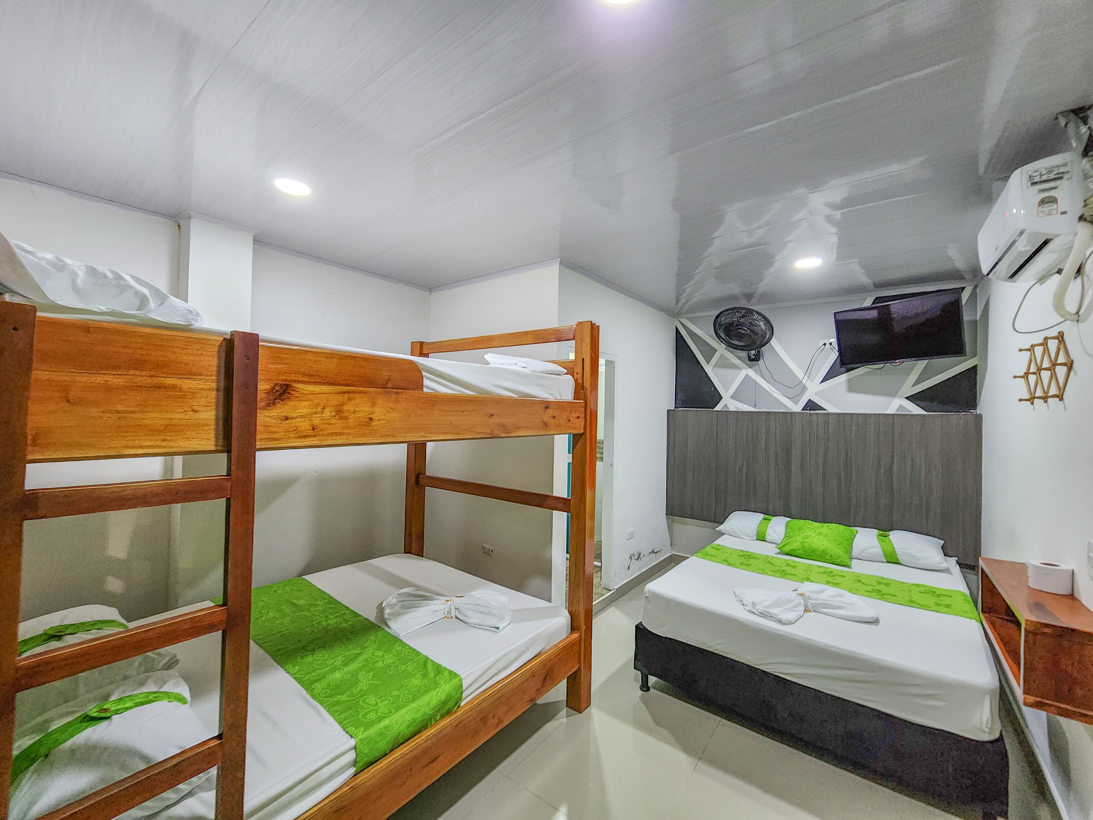 A hotel room with bunk beds, a bed with a green bedspread, a bedside table, a wall-mounted air conditioner, and a TV.