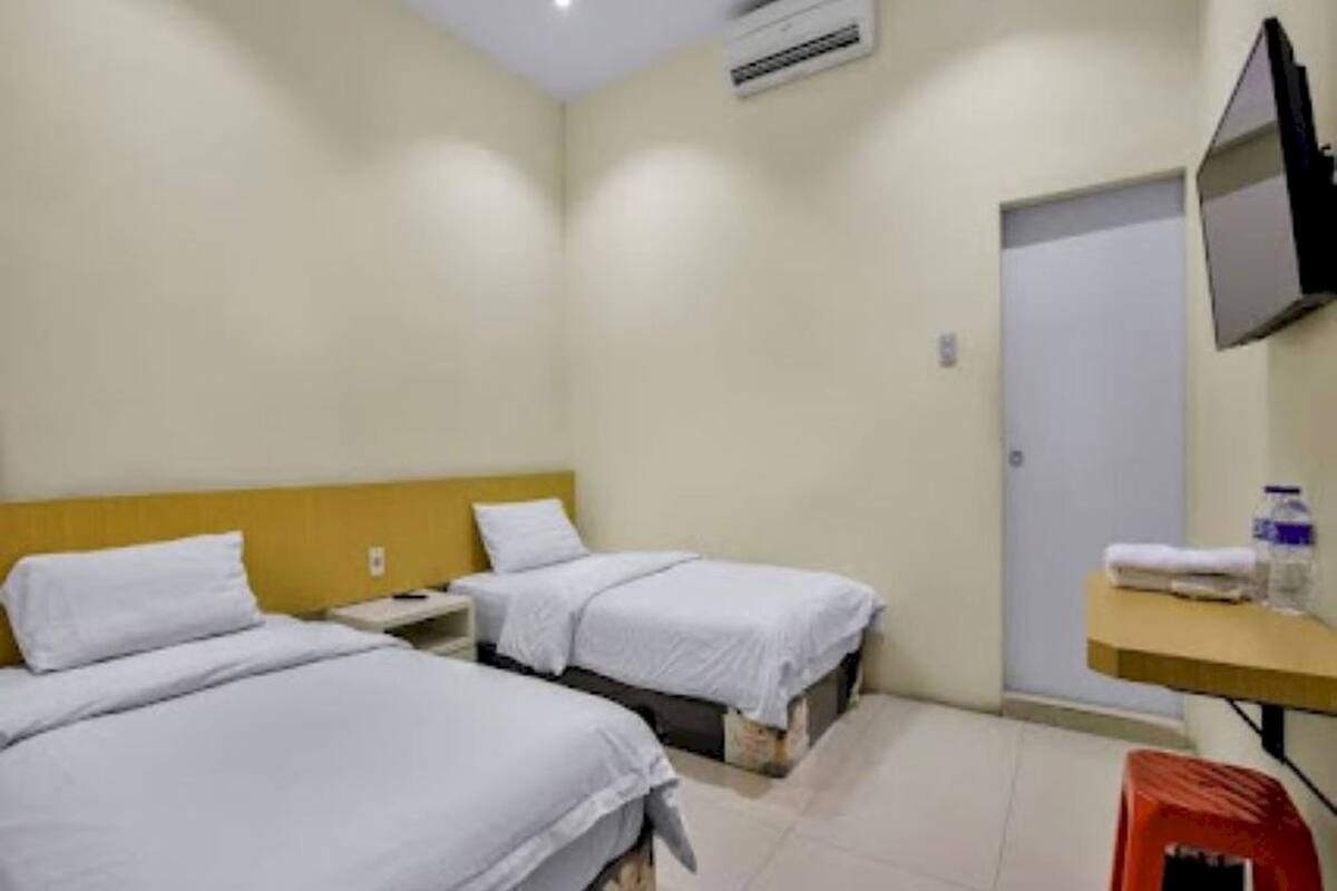 Een hotelkamer met twee bedden, een tv aan de muur, een klein tafeltje met een fles en een handdoek, en een rode kruk.
